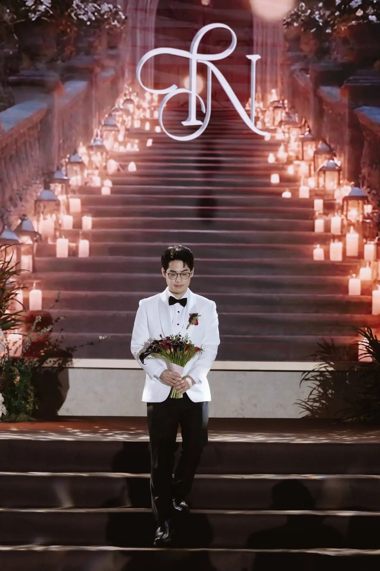 เจ้าบ่าวถือช่อดอกไม้ที่เชิงบันไดซึ่งเรียงรายด้วยเทียนนับร้อย ใต้โมโนแกรมเรืองแสง